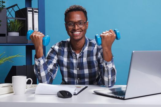 Company athletic businessman lifting weights while sitting at desk in office.