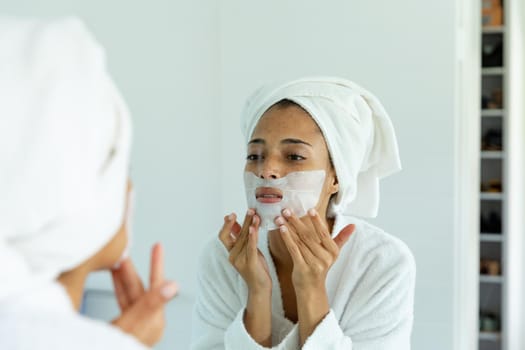 Mixed race woman wearing bathrobe and cleansing face mask in bathroom
