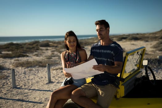 Happy caucasian couple sitting on beach buggy by the sea reading roadmap