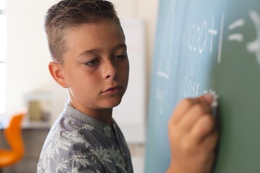 Caucasian boy standing at chalkboard writing in classroom during maths lesson
