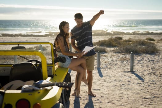 Happy caucasian couple sitting on beach buggy by the sea reading roadmap pointing