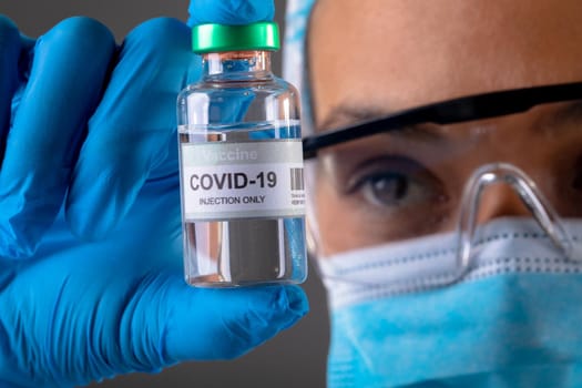 Close up of female surgeon holding covid-19 vaccine bottle against grey background