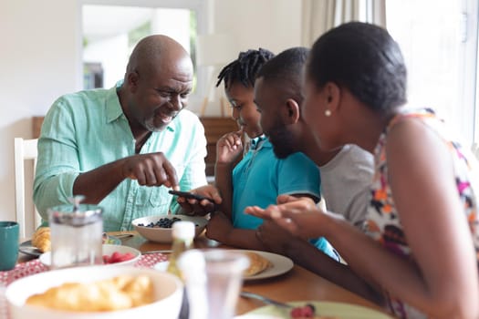 African american grandfather using smartphone with grandson and his parents at table