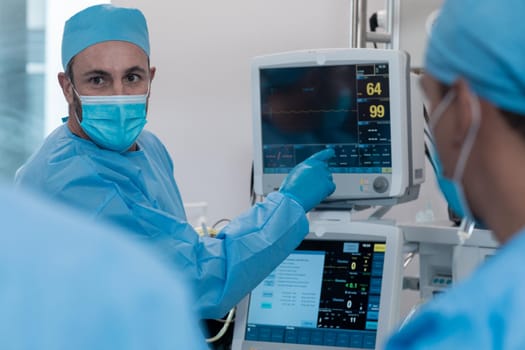 Caucasian male doctor wearing face mask and surgical overalls pointing at monitor