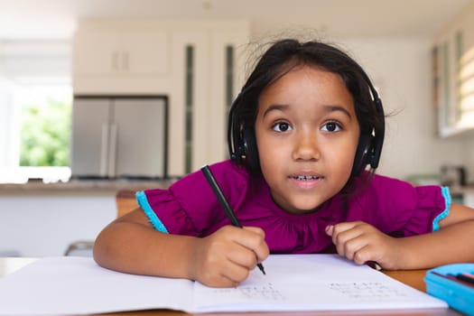 Happy hispanic girl wearing headphones listening and writing during online school lesson