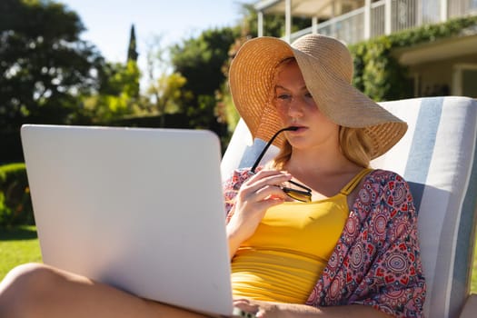 Caucasian woman sitting in sunny garden wearing sunhat using laptop, concentrating