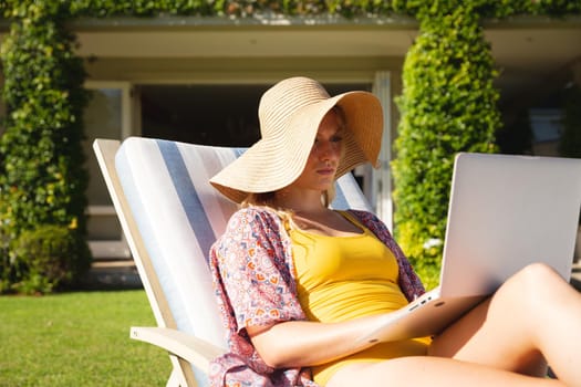 Caucasian woman sitting on sun lounger in sunny garden wearing sunhat using laptop, concentrating