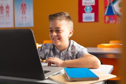Caucasian boy sitting at a desk in classroom using laptop and smiling