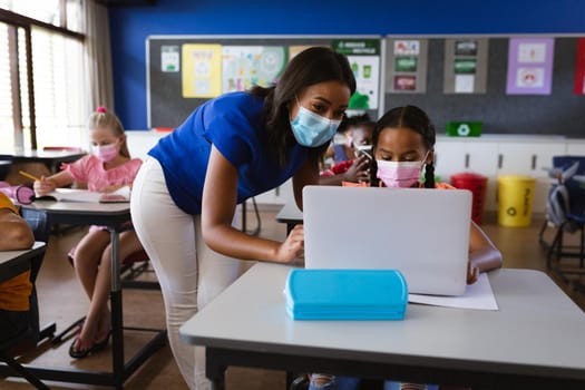 African american female teacher wearing face mask teaching a girl to use laptop at elementary school
