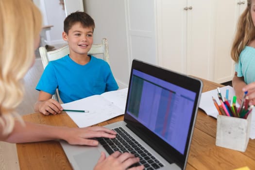 Caucasian mother using laptop and doing homework with her daughter and son smiling in kitchen. family domestic life, spending time working together at home.