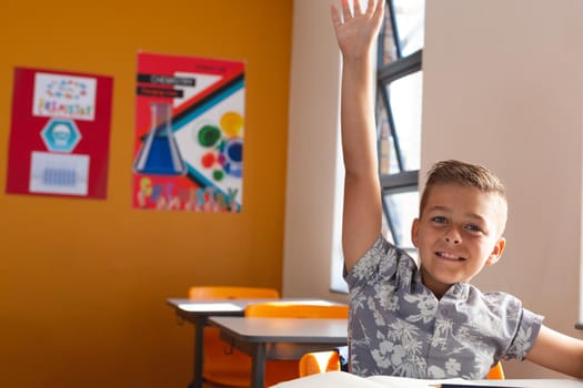 Happy caucasian schoolboy sitting at desk in classroom raising hand during lesson
