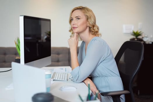 Thoughtful caucasian businesswoman sitting at desk using computer, looking away in thought