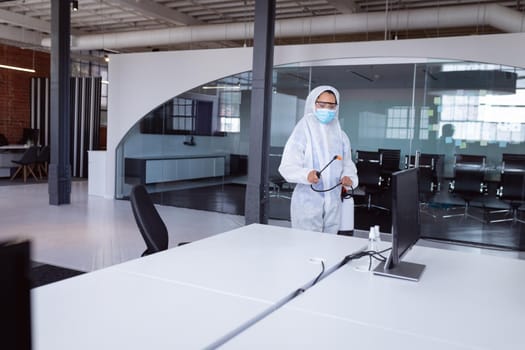 Cleaner wearing ppe suit, glasses and mask disinfecting office workspace