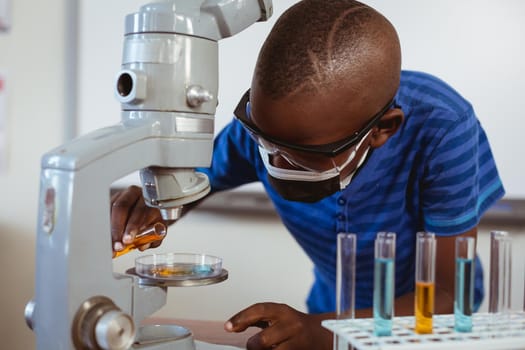 African american schoolboy in safety glasses and face mask using microscope in science class. childhood and education at elementary school during coronavirus covid19 pandemic.