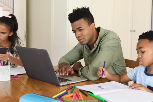 Happy african american father using laptop, daughter and son doing homework at home. family domestic life, spending time learning together at home.