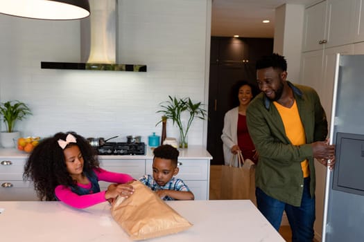 Happy african american siblings unpacking groceries with parents in kitchen