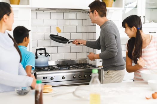 Asian mother, son and daughter watching father tossing pancake for breakfast in kitchen. family enjoying preparing meal together at home.