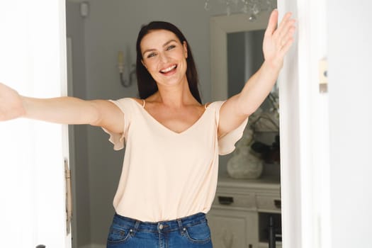 Portrait of happy caucasian woman standing at door and greeting a visitor with open arms