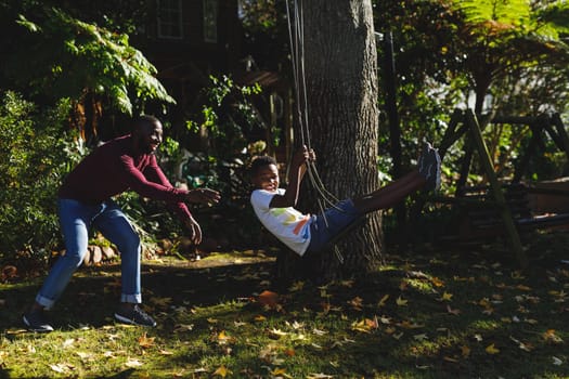 African american father with son having fun and playing on swing in garden. family spending time at home.