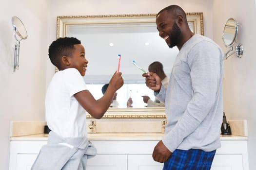 African american father with son having fun and brushing teeth in bathroom. family spending time at home.