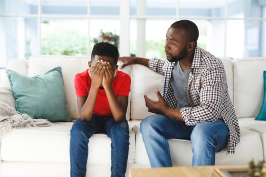 Sad african american son sitting on couch covering face listening to father talking in living room