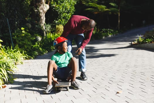 African american father with son smiling and playing with skateboard in garden. family spending time at home.