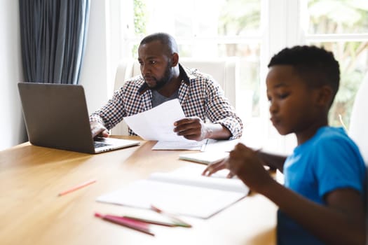 African american father working on laptop in dining room with son sitting with him doing homework