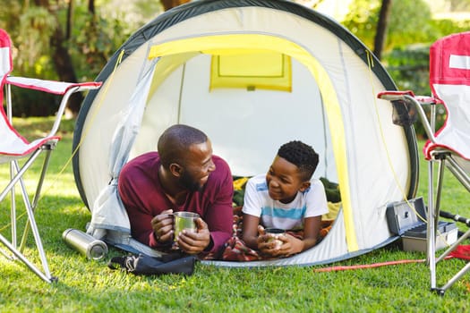 African american father with son having fun and lying in tent in garden. family spending time at home.