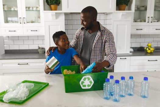 Happy african american father and son in kitchen talking and sorting rubbish for recycling