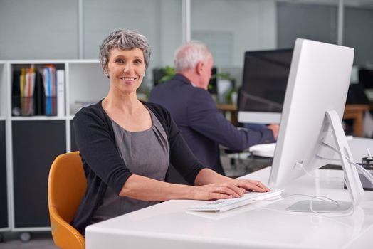 Facing each work day with optimism. Portrait of a smiling mature businesswoman at her desk with a coworker in the background.