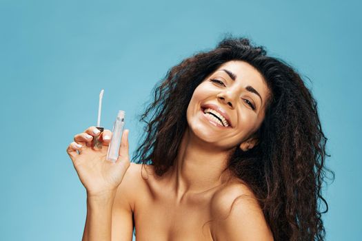 Laughing attractive curly Latin lady hold lip gloss on hand smile look at camera posing isolated over pastel blue background. Cosmetic lips product ad Natural beauty Makeup concept Studio portrait