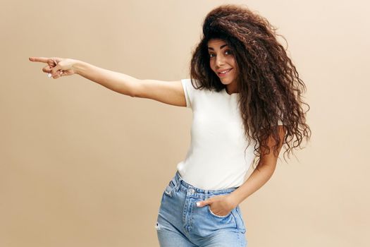SEASONAL SALE OFFER. Excited beautiful Latin female in white t-shirt with afro say Look at this, look at camera, show copy space, free place for design ad. Fashion. Studio shoot over beige background