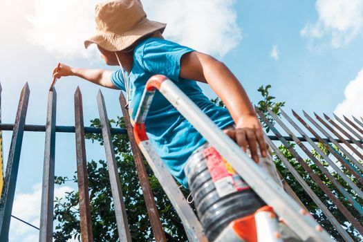 Latin woman dressed in shorts, t-shirt and hat, standing on a ladder standing to paint the highest part of a metal fence