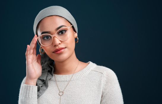 Keeping up with trends. Cropped shot of an attractive young woman standing against a black background alone while wearing spectacles and a headscarf.