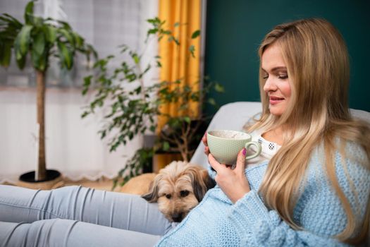 A girl is sitting on a sofa with a cup of coffee and a dog is lying next to her.