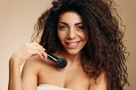 Enjoyed smiling adorable tanned awesome curly Latin lady hold makeup brush on collarbone apply highlighter satisfied posing isolated on pastel beige background look at camera. Cosmetic offer concept