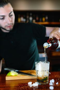 young waiter, pour liquor into a crystal glass filled with ice to prepare a Mojito. Vertical View