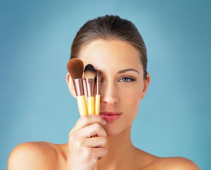 The secret to a flawless makeup routine. Studio portrait of a beautiful young woman holding makeup brushes in front of her eyes against a blue background.