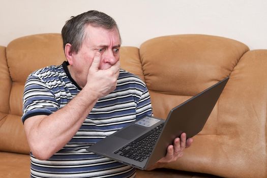 A man with a surprised face holds a laptop in his hands while sitting on couch