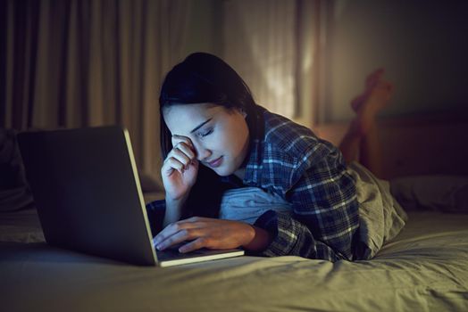 Its been a late night online. Shot of a sleepy young woman using a laptop late at night while lying on her bed in her bedroom.