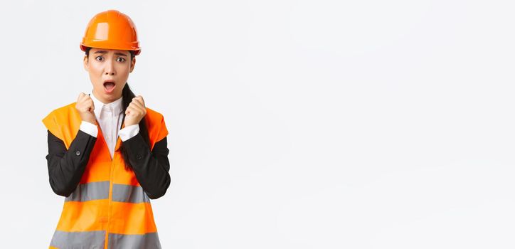 Scared asian female engineer in panic looking shocked and afraid, shivering from fear, wearing safety helmet and reflective jacket, feel concerned and alarmed as having problem at construction zone