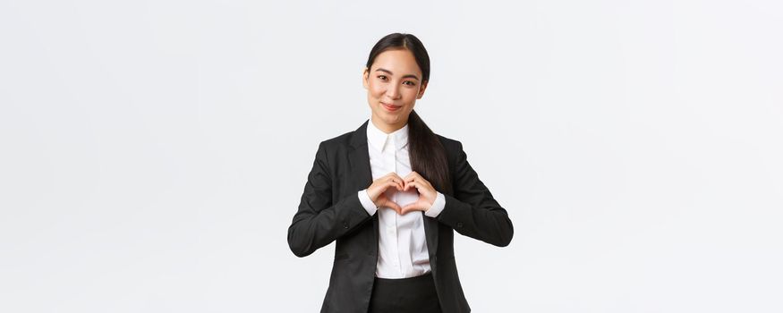 Close-up of pleasant asian businesswoman, saleswoman in black suit care for her clients, showing heart sign and smiling with admiration, standing white background caring and tender