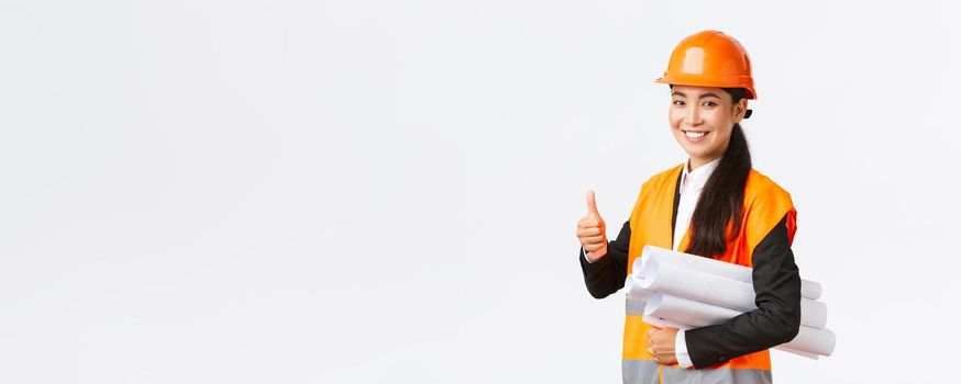 Confident successful female asian construction manager, architect in safety helmet and jacket, showing thumb-up and carry blueprints of building project, guarantee quality, white background