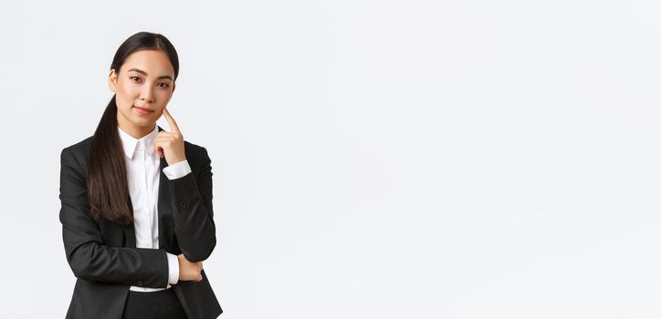 Creative smart asian female manager, saleswoman in suit smiling cunning and looking at camera, have plan, thinking, got excellent idea, standing pleased over white background