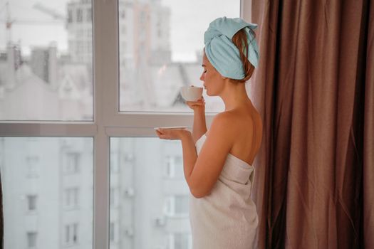 Young serene relaxed woman in spa bath towel drinking hot beverage tea coffee after taking shower bath at home. Beauty treatment, hydration concept