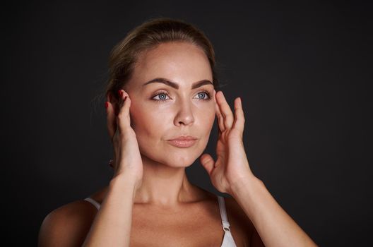 Close-up portrait of an attractive Caucasian woman with fresh glowing skin holding hands on temples, doing massage movements on her face. Anti-aging concept, smoothing, rejuvenating beauty treatment