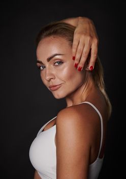 Side portrait of a beautiful confident tanned young Caucasian woman with straight blond hair, natural make-up, beautiful healthy glowing skin posing at the camera on a dark background