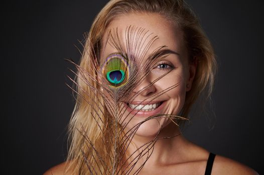 woman, with beautiful healthy glowing skin holding a peacock feather near her face. Black background copy space.