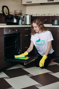 Young woman in yellow protective gloves holding head by hands, looking dirty oven at home kitchen. Brunette girl with curly hair washing oven on kitchen holding hand inside. Housework concept.