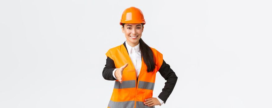 Confident successful female architect, leader of construction in safety helmet, reflective jacket, extand hand for handshake, greeting business partners at building area, standing white background